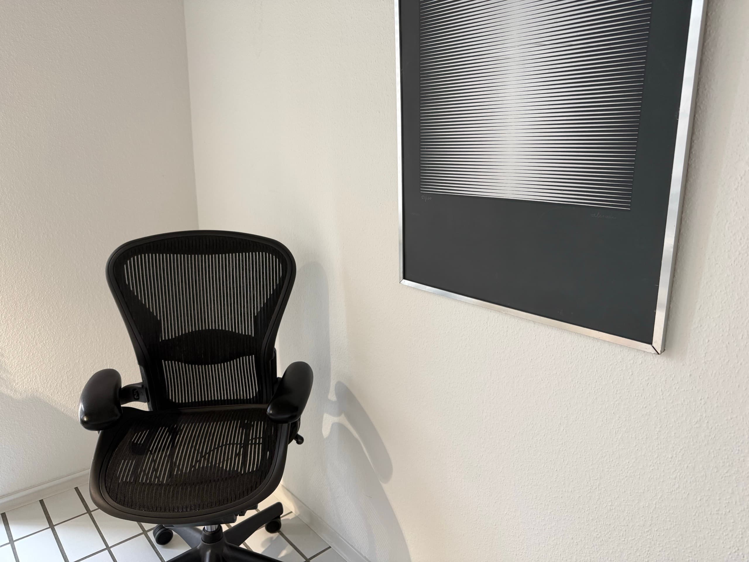 Schwarzer Bürostuhl in einer Zimmerecke neben einer gerahmten Op-Art-Grafik mit Linienmuster